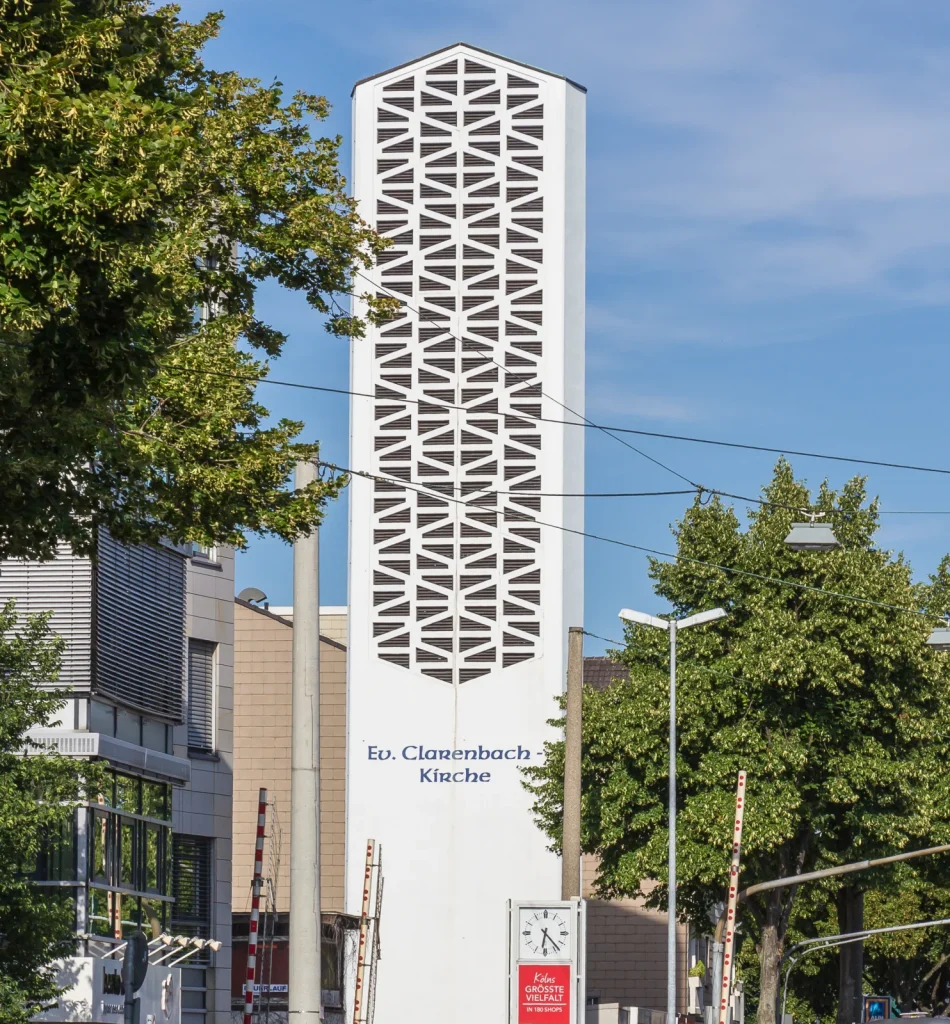 Weißer Glockenturm der Clarenbachkirche mit geometrischen Schallöffnungen vor blauem Himmel.