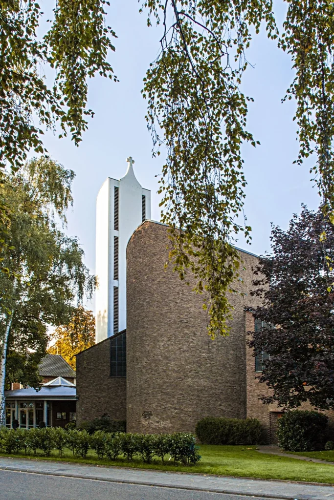 Dietrich-Bonhoeffer-Kirche mit Backsteinwand und Turm hinter Bäumen.