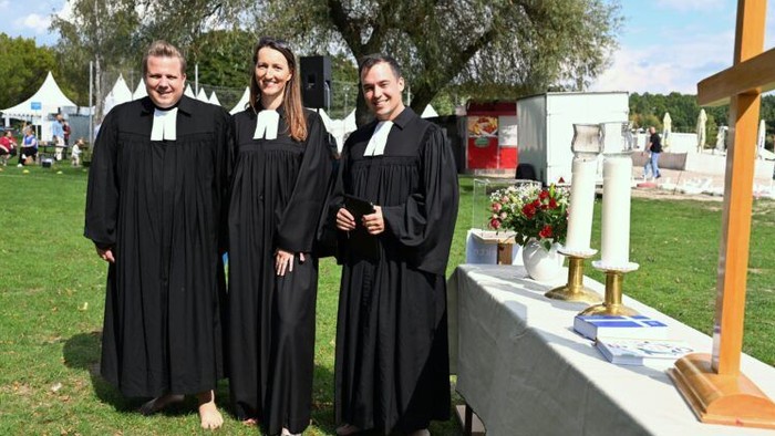 Drei Pfarrpersonen in Talaren neben einem Altar im Freien beim Tauffest am Otto-Maigler-See.